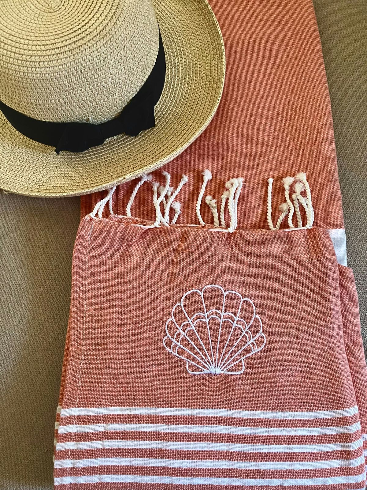 Fouta corail brodée d'un coquillage blanc, posée près d'un chapeau de paille